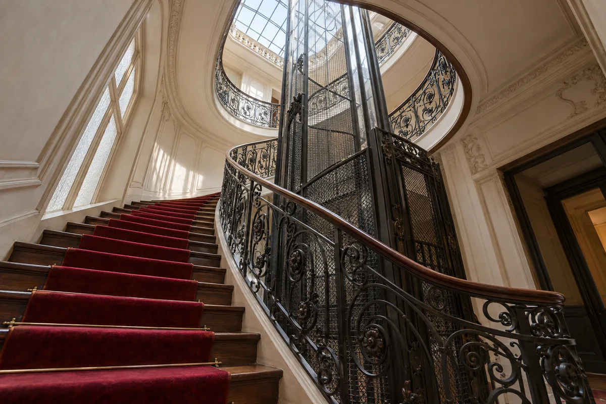 Escalier d’immeuble parisien avec tapis rouge, rampe en fer forgé et ascenseur ancien à grille